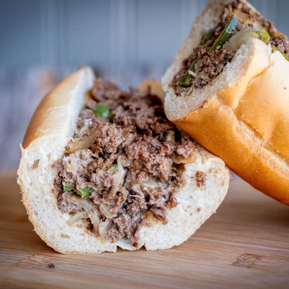 Two halves of a Mandee's Pizza steak and cheese sub with onions and peppers on a wooden board.