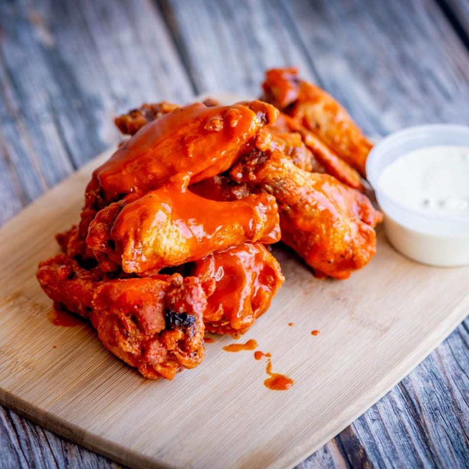 Mandee's Pizza's buffalo wings, coated in red sauce, with a side of ranch dressing on a wooden board.